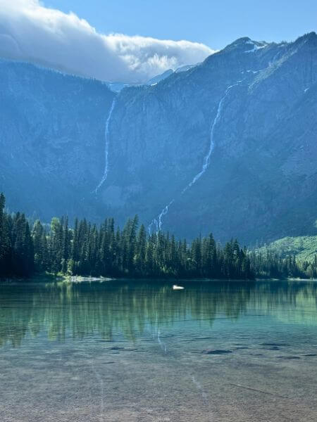 A calm view of Avalanche Lake, surrounded by tall mountains with multiple narrow waterfalls flowing down their cliffs. The crystal-clear water reflects the pine trees lining the shore. The image captures the peaceful beauty of the Avalanche Lake hike.