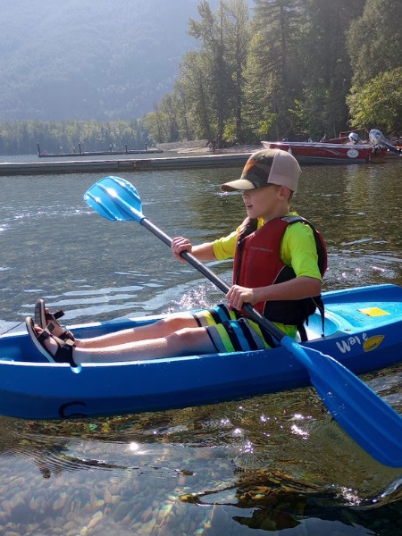 A young boy wearing a life vest paddles a blue kayak on a clear, calm lake. He is focused and smiling as he moves through the water, which reveals smooth rocks beneath the surface. In the background, a dock and boats sit by the shore, surrounded by a dense forest and misty mountains in the distance. The scene captures a peaceful outdoor adventure in nature.