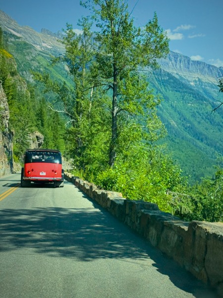 A vintage red tour bus drives along a narrow, scenic mountain road bordered by a low stone barrier. Towering trees and lush green hillsides surround the road, with distant mountains visible in the background under a clear blue sky. The setting conveys a serene and adventurous journey along Going to the Sun Road in Glacier National Park.