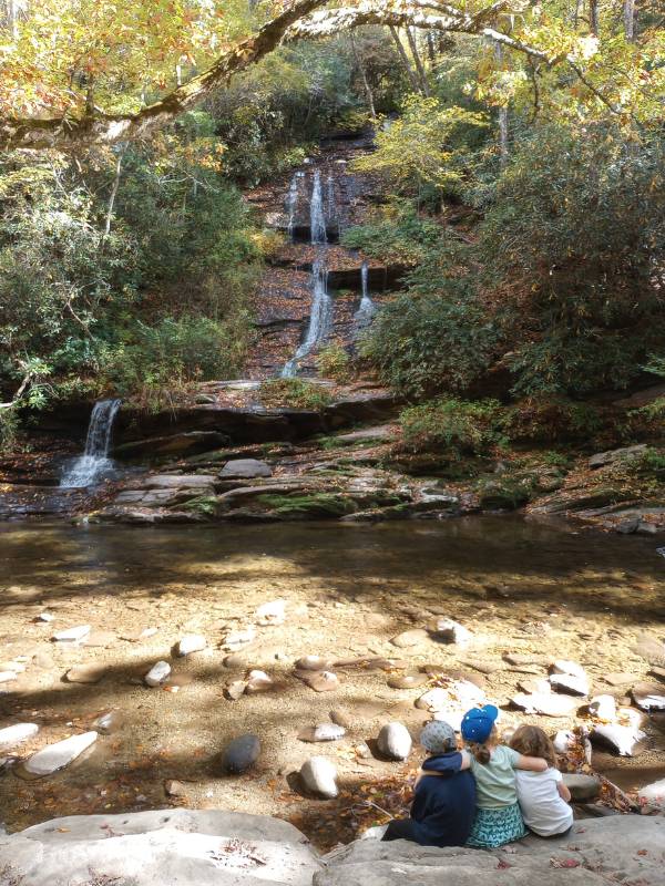 Three children sitting on a rock near a clear stream, admiring a serene waterfall surrounded by lush greenery at Great Smoky Mountains National Park. This peaceful spot highlights the park's natural beauty, a popular destination for family hikes and exploration.