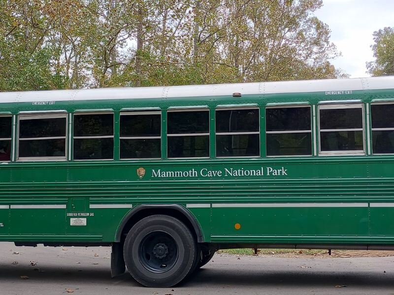 Green shuttle bus with 'Mammoth Cave National Park' written on the side, parked amidst trees in the park. The bus is used for transporting visitors, often requiring reservations for cave tours.