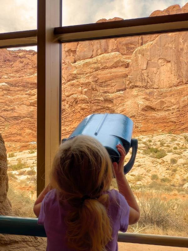 A young girl looking through a telescope at the Arches Visitor Center, gazing out at towering red rock formations through large glass windows. This moment highlights a family-friendly stop during one day in Arches National Park, offering stunning views and interactive experiences for visitors.