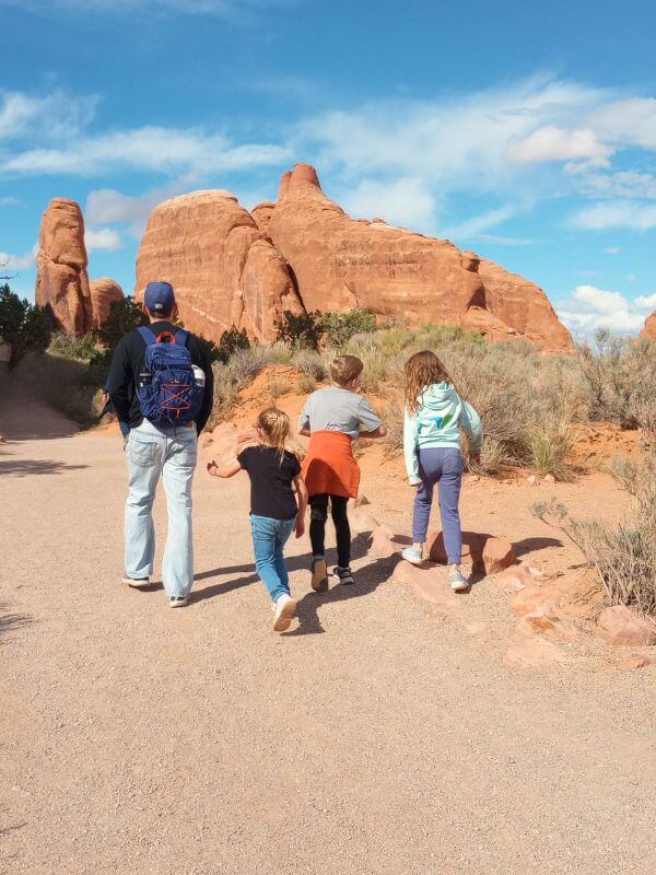 A family hiking together on a sunlit trail in the Devils Garden area of Arches National Park, with red rock formations rising prominently against a bright blue sky. This serene moment captures the adventure of exploring one day in Arches National Park and heading toward iconic spots like Landscape Arch (not pictured).