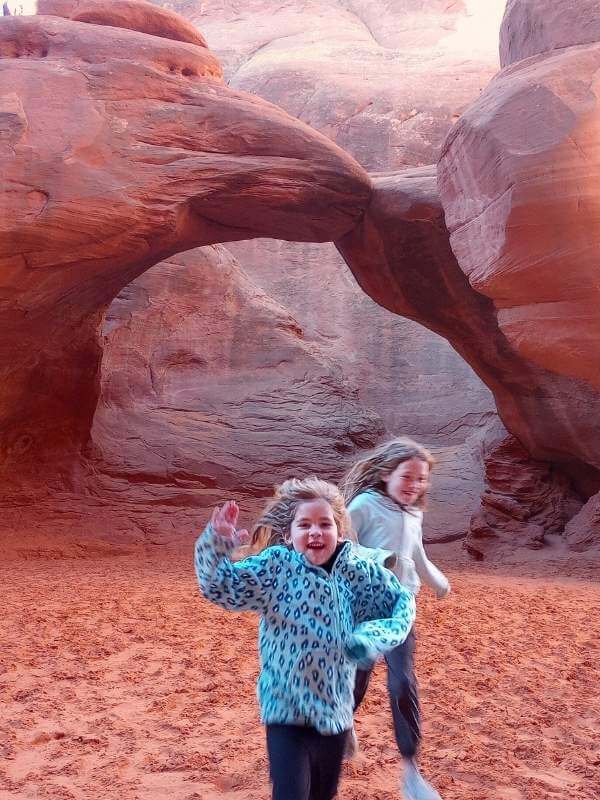 Two children running excitedly on the sandy terrain beneath Sand Dune Arch in Arches National Park, surrounded by striking red rock formations. A perfect snapshot of a family-friendly moment during one day in Arches National Park, highlighting the park's unique landscapes and playful atmosphere.