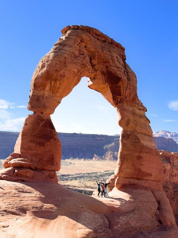 My family standing in front of the red rock formation named Delicate Arch with a clear blue sky behind.