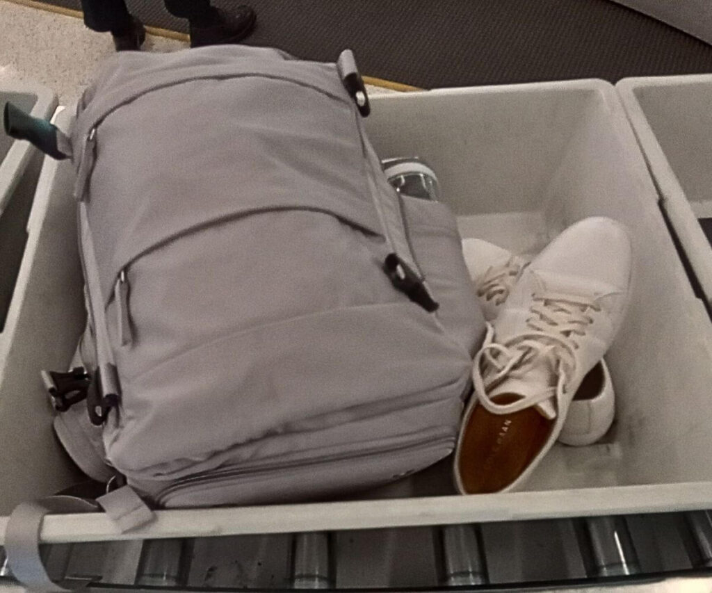 My grey personal item backpack from Coowoz sitting in a screen bin next to a pair of my white shoes.