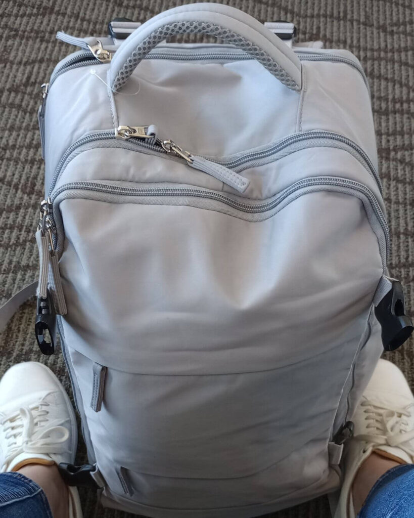 My light grey Coowoz personal item backpack sitting between my feet while I am wearing white sneakers sitting in the airport 